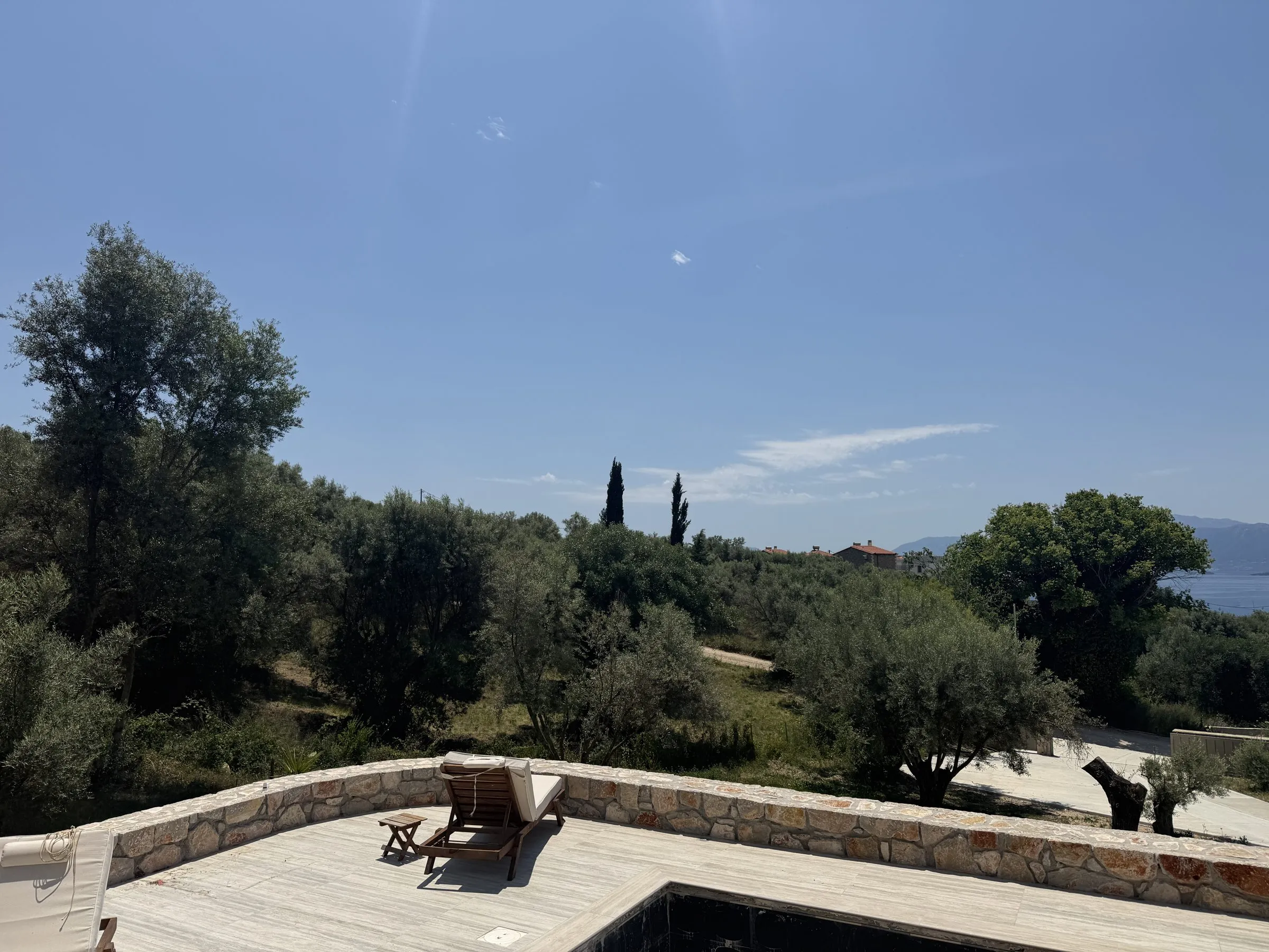 Travertine sun deck at Villa Prima with a wooden lounger, overlooking olive groves and cypress trees toward the Ionian Sea.