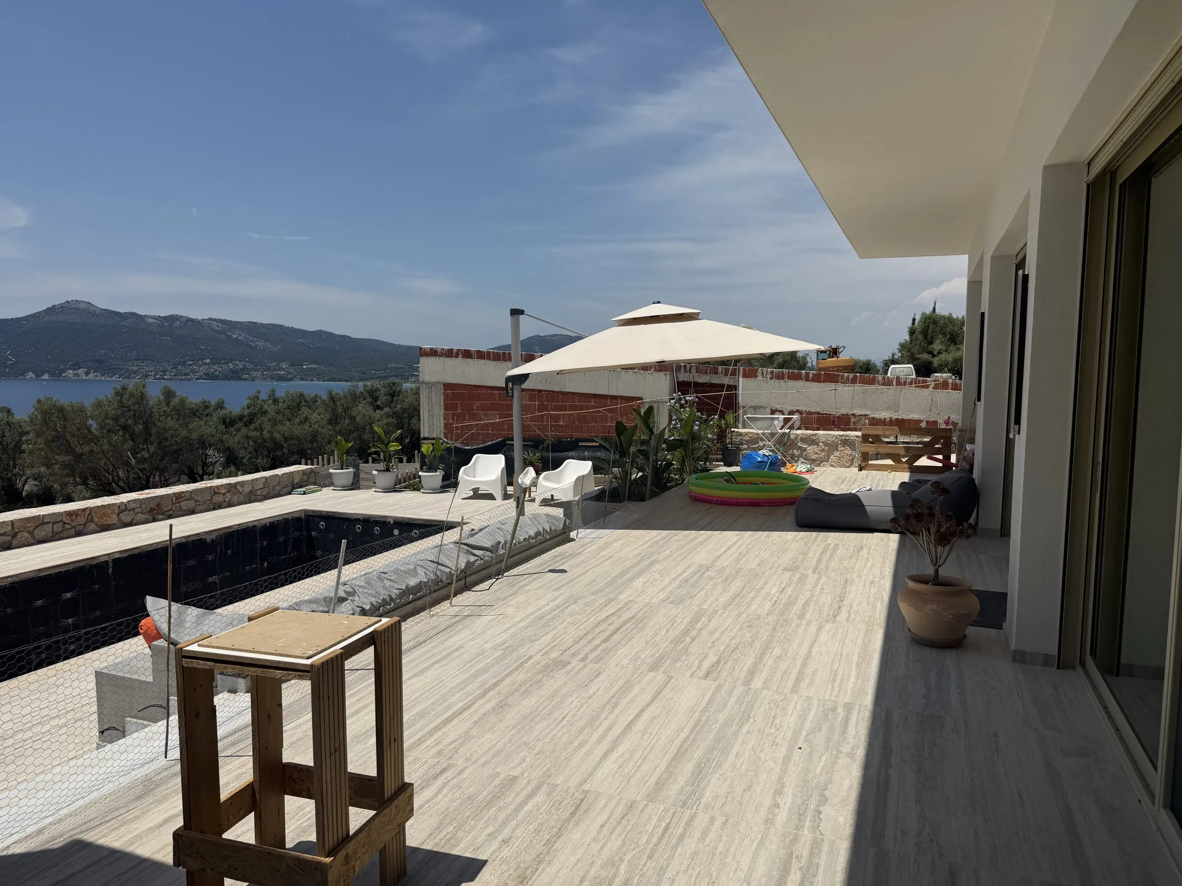 Wide travertine pool terrace at Villa Prima with a parasol and loungers, opening to sea views across Paleros bay.