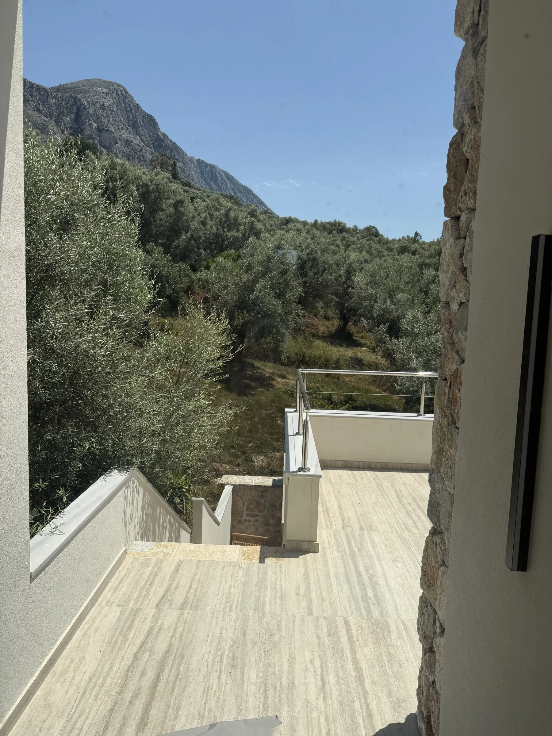 Travertine terrace and external stairs at Villa Prima with a glass-and-steel railing, framed by olive trees and the mountain.