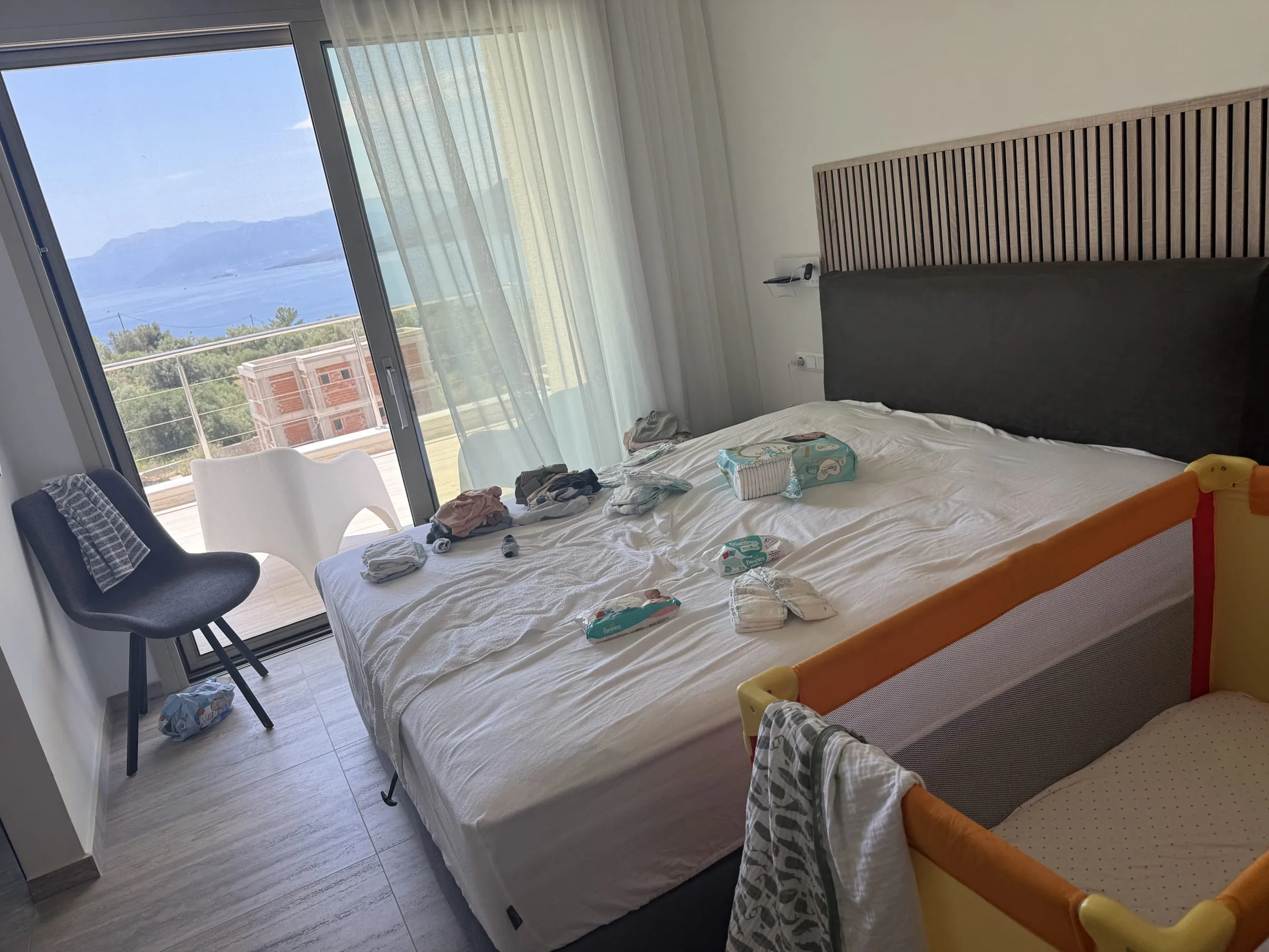 Family bedroom at Villa Prima with a large bed, slatted timber headboard, travel cot and a balcony framing sea and mountain views.