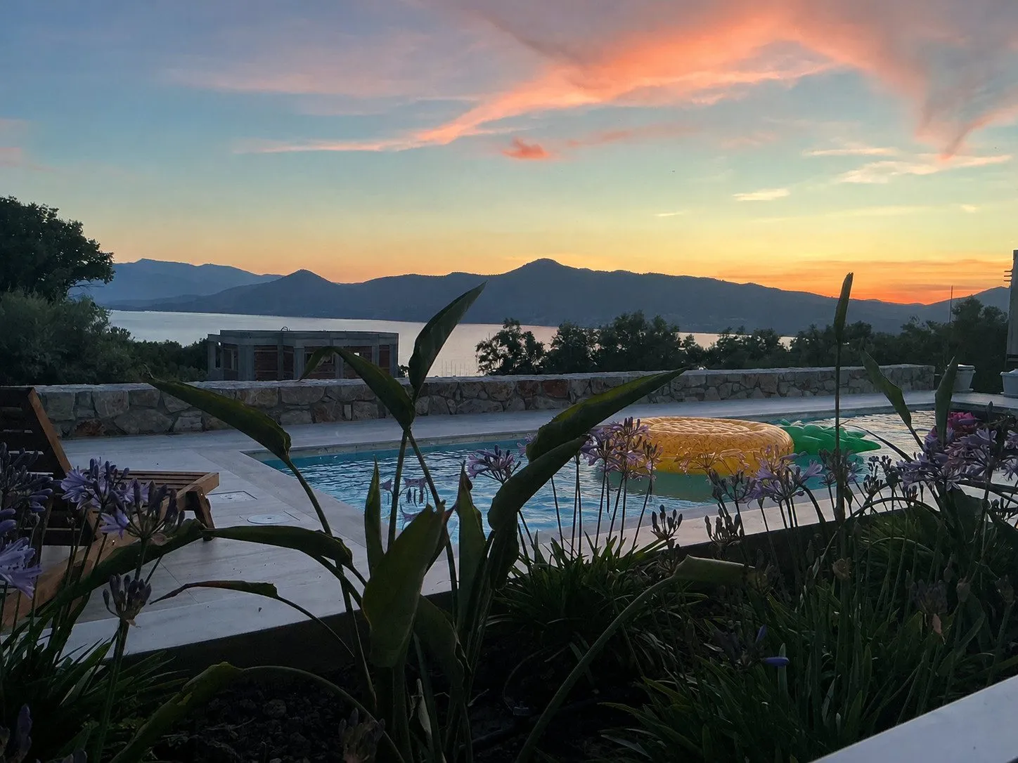 The pool terrace at Villa Prima at sunset, agapanthus flowers in the foreground and the Ionian bay and mountains glowing pink and gold beyond.
