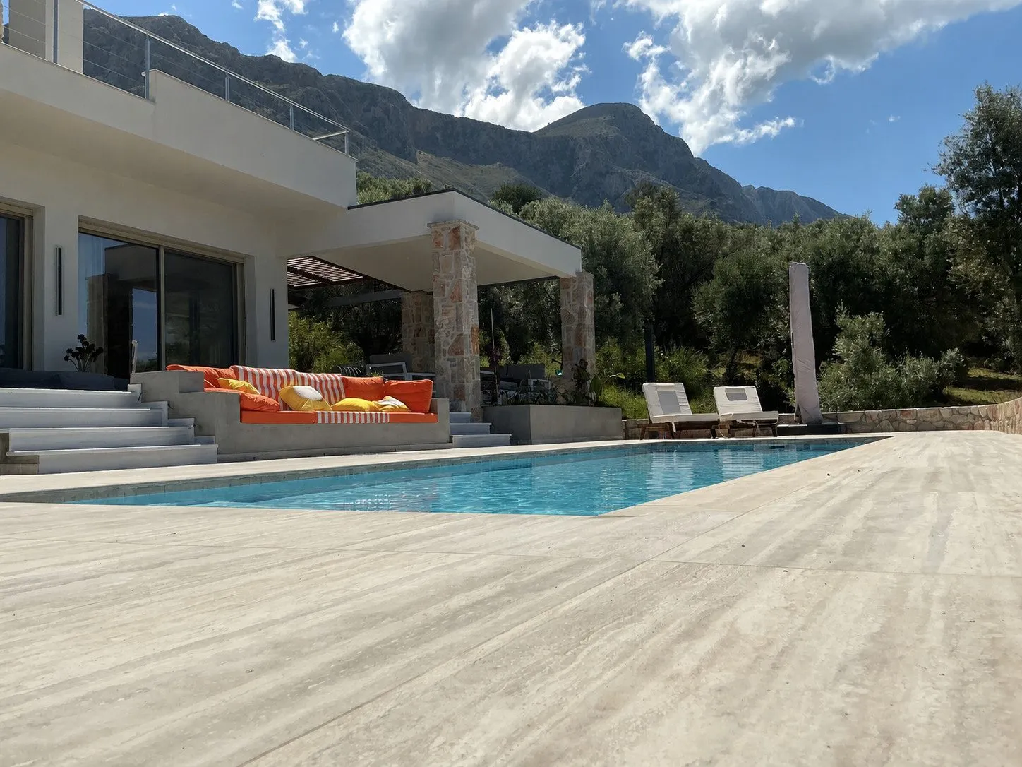 The travertine pool deck at Villa Prima with the white villa, a shaded terrace lounge and the limestone mountain rising behind under a blue sky.